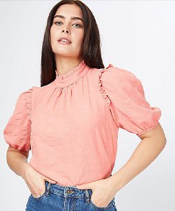 Woman with dark brown hair poses with hands in pockets wearing coral seersucker high neck frill blouse and jeans.