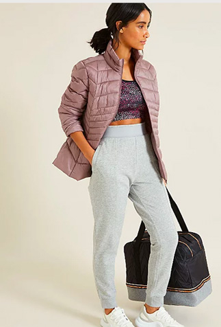 Woman holding a bag wearing a pink jacket over a sports bra, grey joggers and white trainers