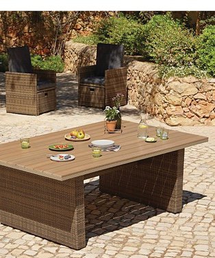 A large outdoor table on a patio with crockery, food and decorative vases on top, with matching outdoor armchairs nearby