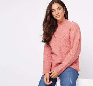 Woman sitting on a white box wearing a pink diamond knit jumper and distressed jeans