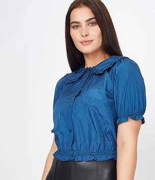 Woman poses wearing blue taffeta frill collar boxy blouse and black faux leather trousers.