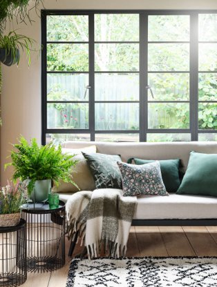 A sofa topped with a selection of green cushions and throws next to side tables with plants and vases, all in front of a bright window.