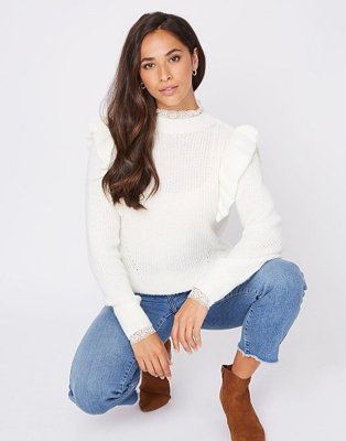 A woman crouching wearing blue jeans with frayed edges, brown boots and a cream ribbed lace collar ruffle jumper