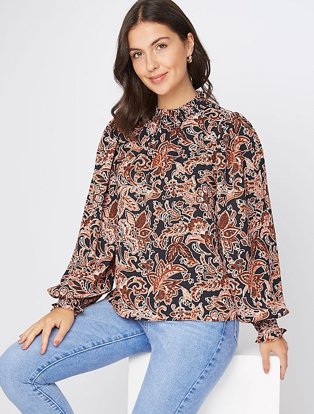 Brunette woman sits on white box wearing burnt orange paisley print high neck blouse and light blue jeans.