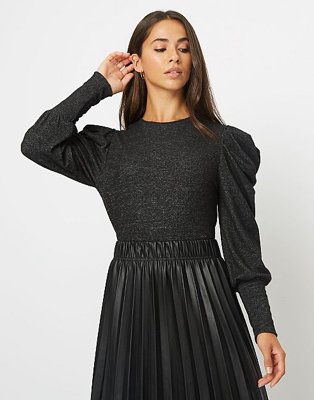 Brunette woman poses raising left hand to face wearing black pleated sleeve jumper and black PU pleated skirt.