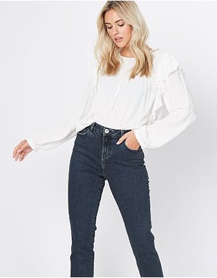 Blonde woman poses wearing white textured high neck frill detail blouse and dark blue jeans.