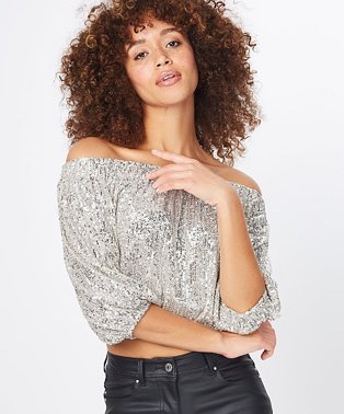 Posing woman wearing a silver effect sequinned cropped bardot blouse with black jeans.