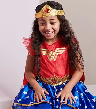 Girl poses sitting down smiling with hands on knees wearing DC Comics Wonder Woman Fancy Dress Costume.