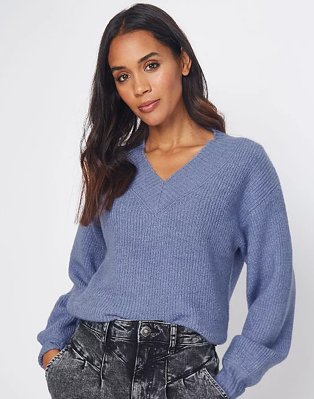 Woman with long brunette hair poses with hands in pockets wearing blue volume sleeve ribbed jumper and acid wash jeans.