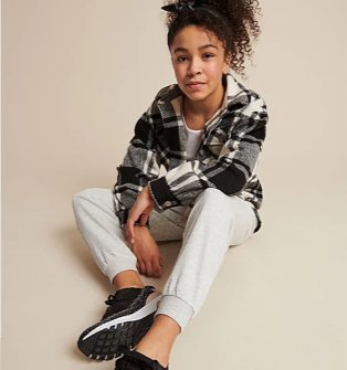 Girl sits on floor wearing a black, white and grey checkered shirt, grey joggers and black pumps.