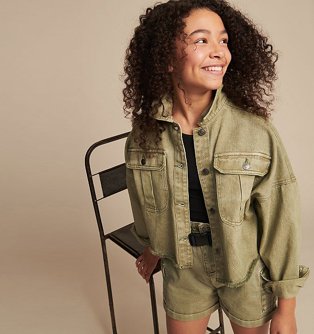 Girl stands in front of chair smiling wearing a black top, khaki jacket and shorts with black belt.