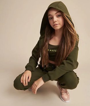 Girl crouches down wearing a khaki slogan top, hoodie and joggers.