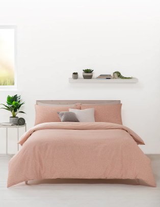 Double bed with pink duvet set and grey and cream scatter cushions with bedside table and artificial plant to one side and white shelving in the background.
