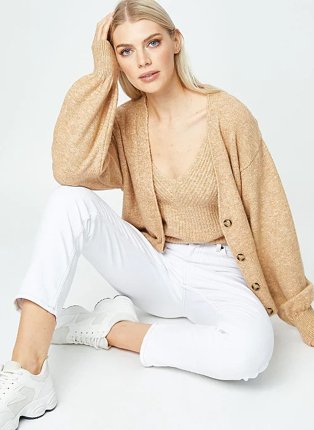 Woman poses sitting on the floor wearing camel knitted vest top, camel knitted cardigan, white jeans and white trainers.
