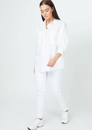 Woman looks to the floor posing with one foot forward wearing white shirt, white jeans and white trainers.