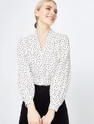 Woman looks over shoulder smiling with hands in front of her wearing white and black polka blouse and black jeans.