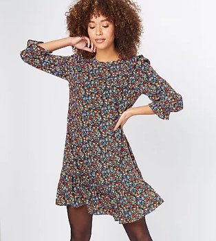 Woman poses with one hand to side of face and one hand on hip wearing multicoloured tiered hem floral dress and black tights.