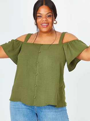 Woman poses smiling with arms raised wearing khaki cold shoulder top and blue mid-wash jeans.