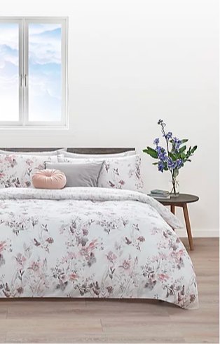 Double bed features floral duvet set with grey and pink scatter cushions with wooden side table and artificial plant.