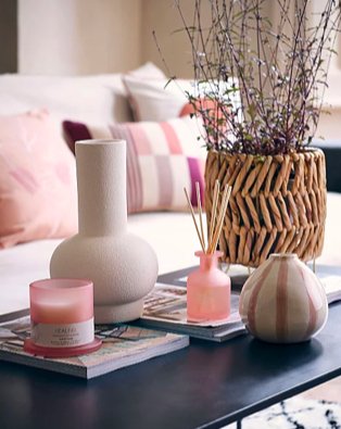 Coffee table features cream vase, artificial plant in wicker vase, pink candle, pink reed diffuser and pink and cream striped vase with cream sofa in the background.