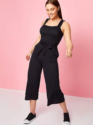 Woman poses smiling wearing black tie waist jumpsuit and black contrast toe pumps.