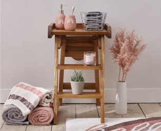 Wooden ladder table features pink rainbow print dispenser and tumbler set, grey hand towels in wire basket, pink candle and artificial potted plant with selection of grey, pink and cream towels and artificial plant on floor.