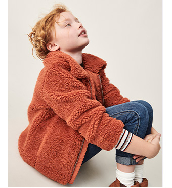 Child sitting down wearing a brown borg coat and jeans 