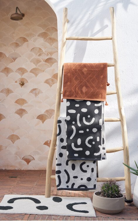 A selection of Sahara towels draped on a ladder, an abstract print bathmat and a plant