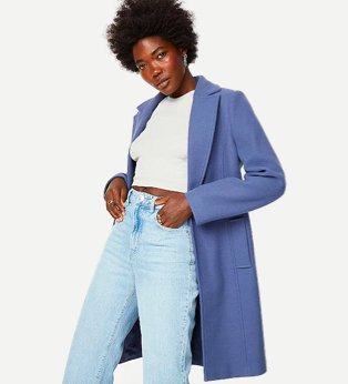 Woman poses with hands in pockets wearing white knitted crop top, light blue high-rise jeans and blue one button longline Crombie coat.