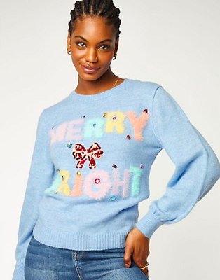 Woman poses wearing blue Christmas jumper and mid-blue jeans.