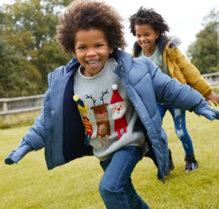 Two smiling children run in field wearing Christmas jumpers, padded coats and blue jeans.
