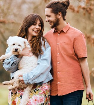 Woman smiles holding white dog wearing pink floral dress and light blue denim jacket looking up at man wearing orange shirt and blue jeans.