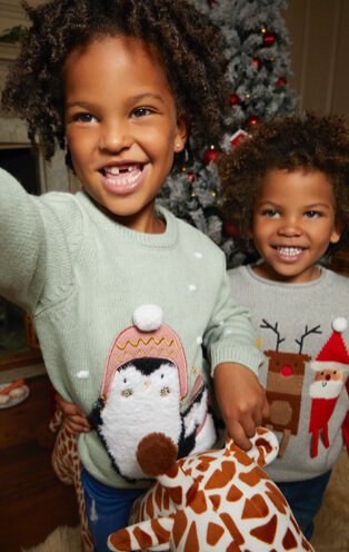 Two children huddle together smiling wearing knitted Christmas jumpers with Christmas tree in the backrgound.