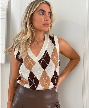 Woman poses with hand on hip wearing cream and brown G21 argyle knitted sweater vest tucked into chocolate brown faux leather trousers.