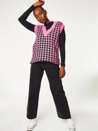 Woman poses with hand to head wearing black long-sleeved top under G21 pink houndstooth longline knitted sweater vest.
