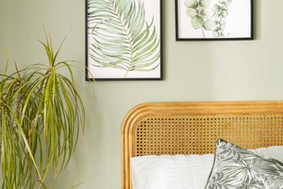 Room features leaf wall prints, double bed with wicker headboard and large artificial plant.