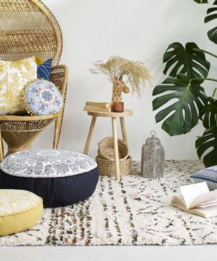 Room features wicker basket chair topped with blue and yellow printed cushions with wooden side table topped with giraffe vase, books and a candle with large artificial plant in corner of room.