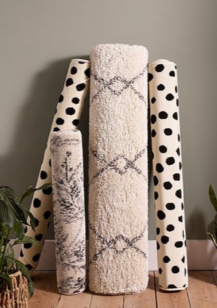 Assortment of abstract printed rugs stood upright on wooden floor against grey wall.