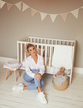 Billie Faiers sitting on the floor in front of a white wooden crib with baby bedding, toys and cushions surrounding her