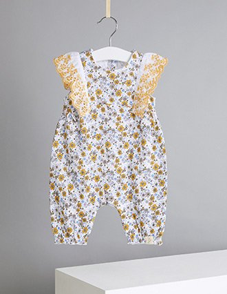 An ochre floral broderie anglaise trim romper hanging from a white hanger on a ribbon