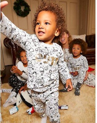 Young boy smiles wearing Peanuts™ Snoopy Slogan family Christmas pyjamas with woman and two young boys huddled together in the background.