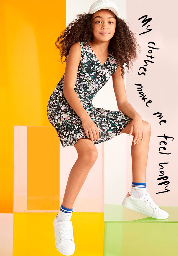 Girl sits on box wearing green floral dress, white baseball cap, stripey socks and white trainers.