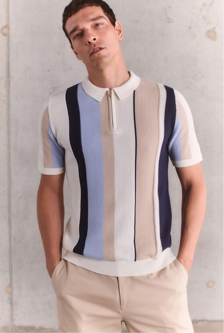 Man poses with hands in pockets wearing a striped polo shirt and cream chinos.