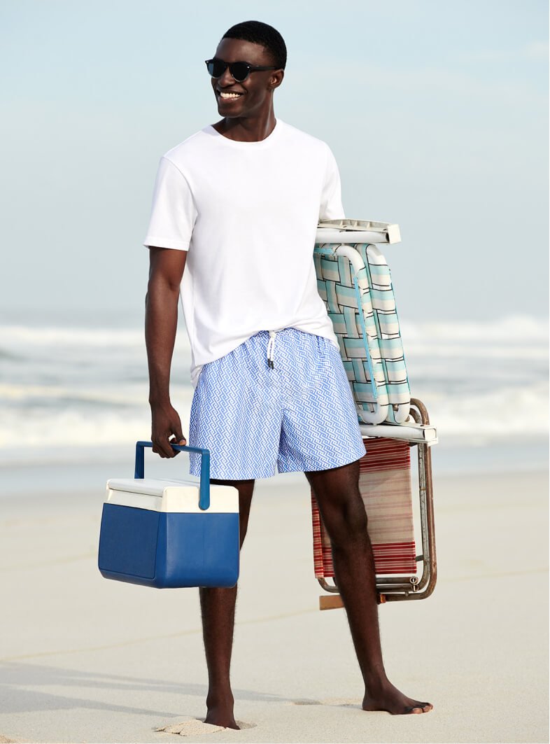 Man on beach holding cooler box and chairs wearing white t-shirt and blue and white printed shorts.