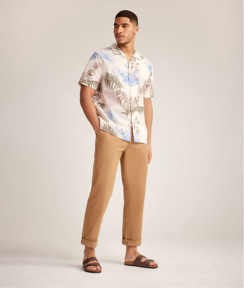 Man poses with one hand in pocket wearing a palm print shirt, tan chinos and brown sliders.