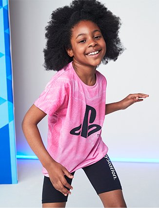 Girl poses smiling wearing pink PlayStation logo t-shirt and black shorts with white PlayStation lettering.