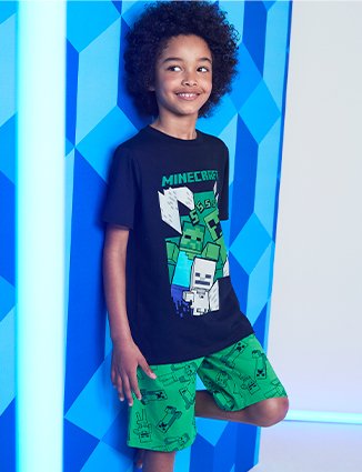 Boy poses with one leg against a wall wearing black Minecraft t-shirt and matching green Minecraft shorts.