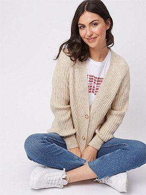 Woman sitting in a white slogan T-shirt, pink cardigan, jeans and white trainers