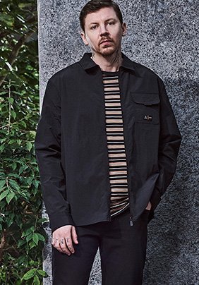 Professor Green stands with one hand in pocket wearing blue and cream striped t-shirt, black collared jacket and black jeans.