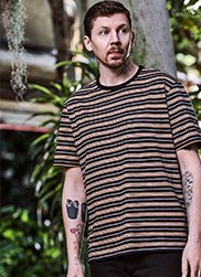 Professor Green poses with greenery in the background wearing blue and cream striped t-shirt.